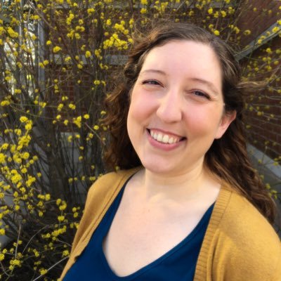 Anna is a white woman with long, curly, brown hair who is smiling and looking at the camera. She is wearing a dark blue top and mustard-yellow cardigan which matches the yellow flowers on the blooming plants behind her.