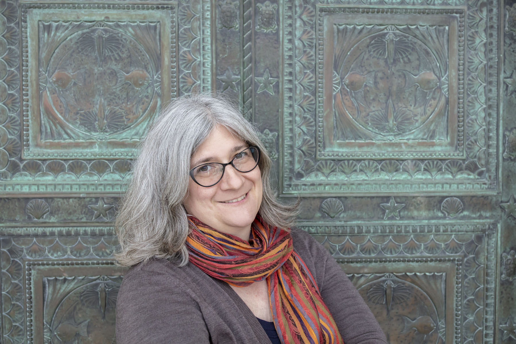 Kris is a white woman with shoulder length gray hair, smiling as she stands in front of a patterned door.