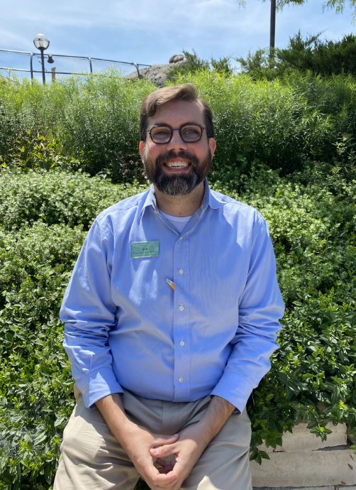 Bill is a white man in his 40’s with short, dark hair and beard, wearing round glasses. He is pictured in a blue dress shirt seated in front of green leafy plants at the zoo and smiling to camera.