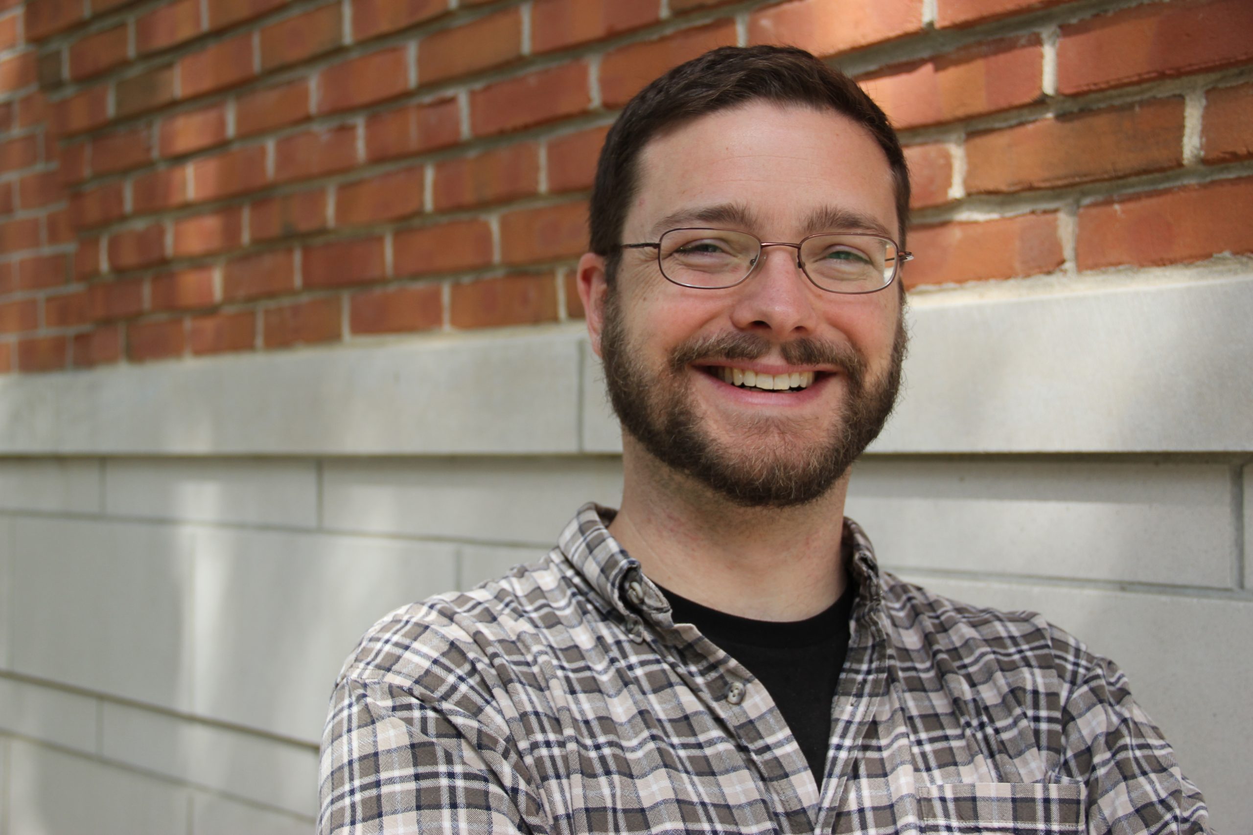 Matt is a white man with short brown hair, a close shaven beard, and a broad smile. He wears glasses and a light brown plaid shirt, and stands in front of a red brick wall.